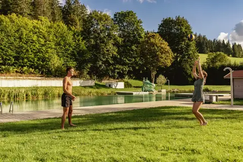 Paar spielt Volleyball auf Wiese am Naturbadeteich, im Hintergrund Rutsche und Wald