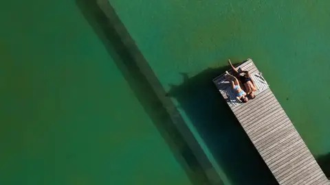 Luftaufnahme: zwei Personen liegen auf einem Holzsteg im türkisgrünen Wasser