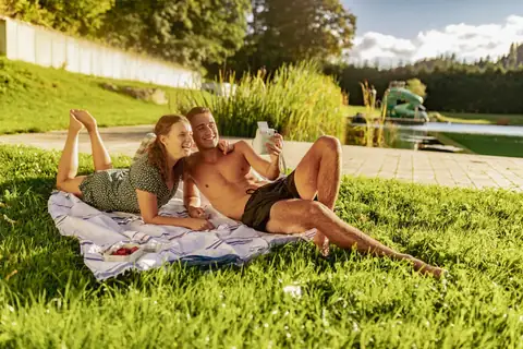 Junges Paar liegt lachend auf einer Picknickdecke im Gras vor einem Natursee mit Steg