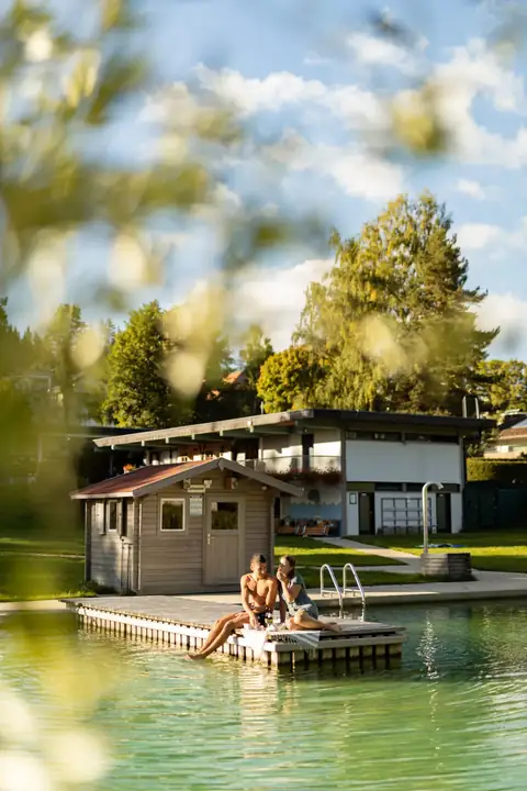 Paar sitzt entspannt am Steg des Naturfreibads mit Blick auf Wasser, Badehaus und Wiese