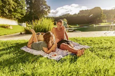 Junges Paar liegt auf Picknickdecke am Grasufer eines Naturbadesees mit Steg und Wald im Hintergrund