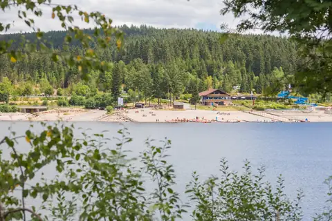 Schluchsee mit Sandstrand, Bootsverleih und Gastronomiegebäude, umgeben von Schwarzwaldtannen