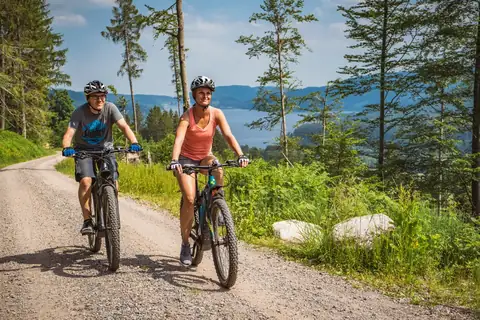 Zwei Radfahrer mit Helm auf Schotterweg im Wald, im Hintergrund der Schluchsee und bewaldete Hügel