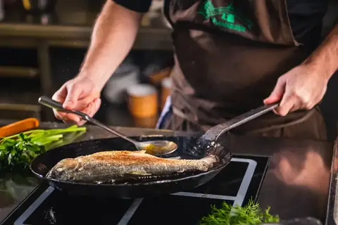 Koch brät ganzen Fisch in Pfanne, Nahaufnahme von Händen mit Kelle und Löffel
