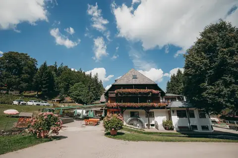 Schwarzwaldgasthaus mit Fachwerkbalkon, Blumenschmuck und Wasserrad vor Baumkulisse und blauem Himmel
