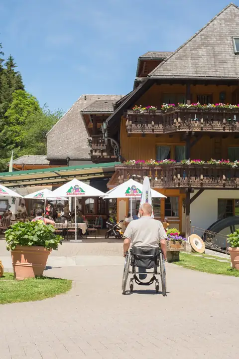 Schwarzwaldhaus mit Balkonen und Blumenschmuck, Terrasse mit Sonnenschirmen, Mann im Rollstuhl auf dem Weg