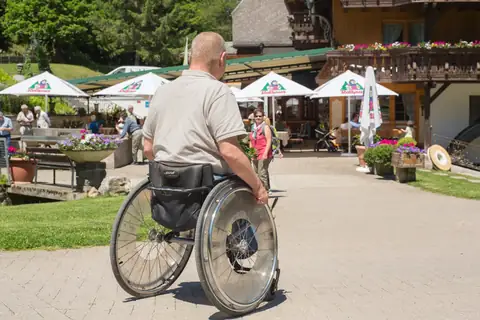 Rollstuhlfahrer vor Schwarzwaldgasthaus mit Sonnenschirmen und Gästen auf der Terrasse