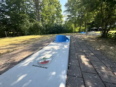Weiße Minigolfbahn mit blauem Hindernis, ringsum Wiese und Laubbäume