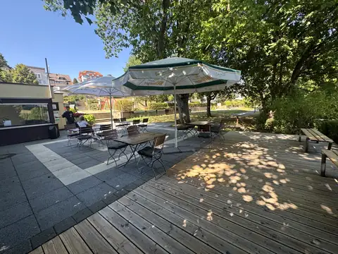 Terrasse des Minigolfcafés mit Holzdeck, Sonnenschirmen und Bistrotischen unter schattigen Bäumen