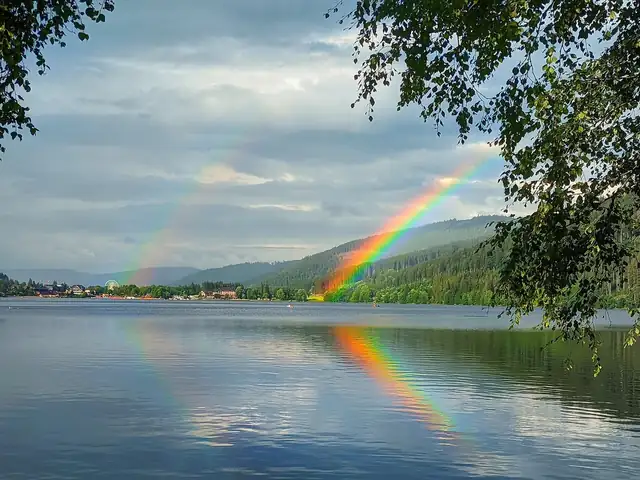 Doppelregenbogen über dem Titisee mit Spiegelung im Wasser, Riesenrad und Wald im Hintergrund