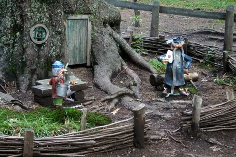 Zwei Wichtelfiguren aus bemaltem Metall vor einem Baum mit kleiner Tür, umgeben von Flechtwerk