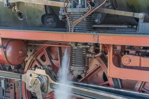 Nahaufnahme von Zylinder, Rohrleitungen und Rad einer Dampflokomotive mit austretendem Dampf