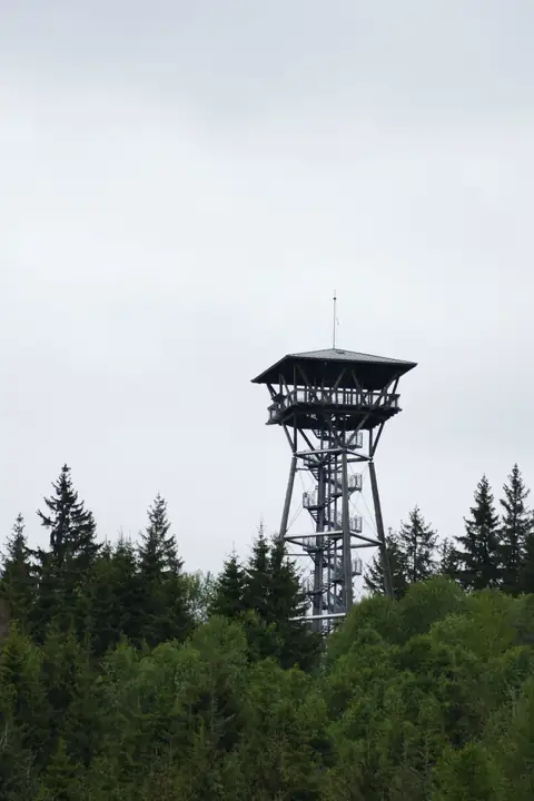 Holzturm mit spiralförmiger Treppe und Aussichtsplattform über Waldkulisse, bewölkter Himmel
