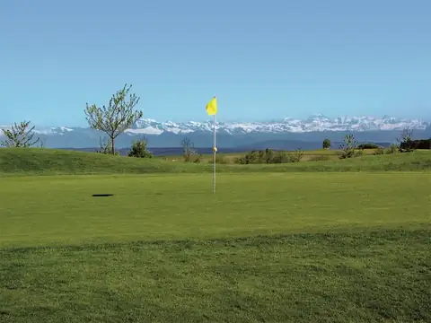 Golfgrün mit gelber Fahne auf dem Hochplateau, am Horizont die schneebedeckte Alpenkette