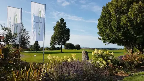 Blühende Staudenbeete mit Hortensien und Lavendel vor den Clubfahnen, dahinter Fairway und Findling