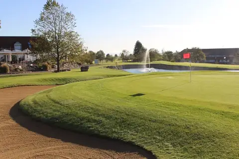 Golfgrün mit roter Fahne hinter einem Sandbunker, dahinter der Teich mit Fontäne und das Clubhaus des Golfclubs Obere Alp