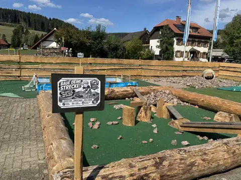 Bereich zum Selberbauen mit Schild „Baue deine Strecke!“, Holzstämmen, Steinen und Kunstrasen, im Hintergrund Schwarzwaldhäuser