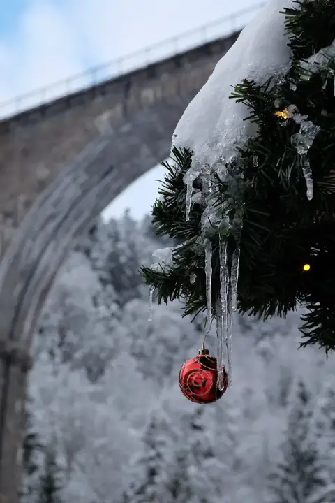 Roter Christbaumschmuck mit Eiszapfen am Tannenzweig, im Hintergrund verschneites Viadukt
