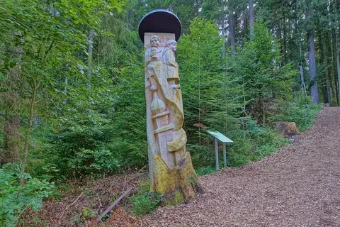 Geschnitzte Holzstele mit zwei Figuren am Wegrand im Wald, Infotafel im Hintergrund