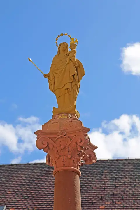 Vergoldete Mariensäule mit Kind auf Steinsockel vor Kirchendach und blauem Himmel