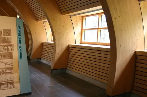 Holzgewölbe-Innenraum mit gebogenen Holzrippen, Sitzbank und Ausstellungstafel 'Mensch und Natur'
