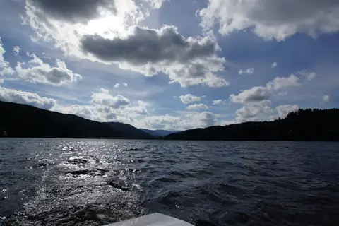 Blick vom Boot auf den Titisee mit bewaldeten Schwarzwaldhügeln und dramatischem Wolkenhimmel