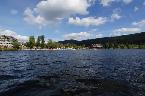 Blick vom Titisee auf Seepromenade mit Bootsstegen, Hotels und bewaldeten Hügeln im Hintergrund